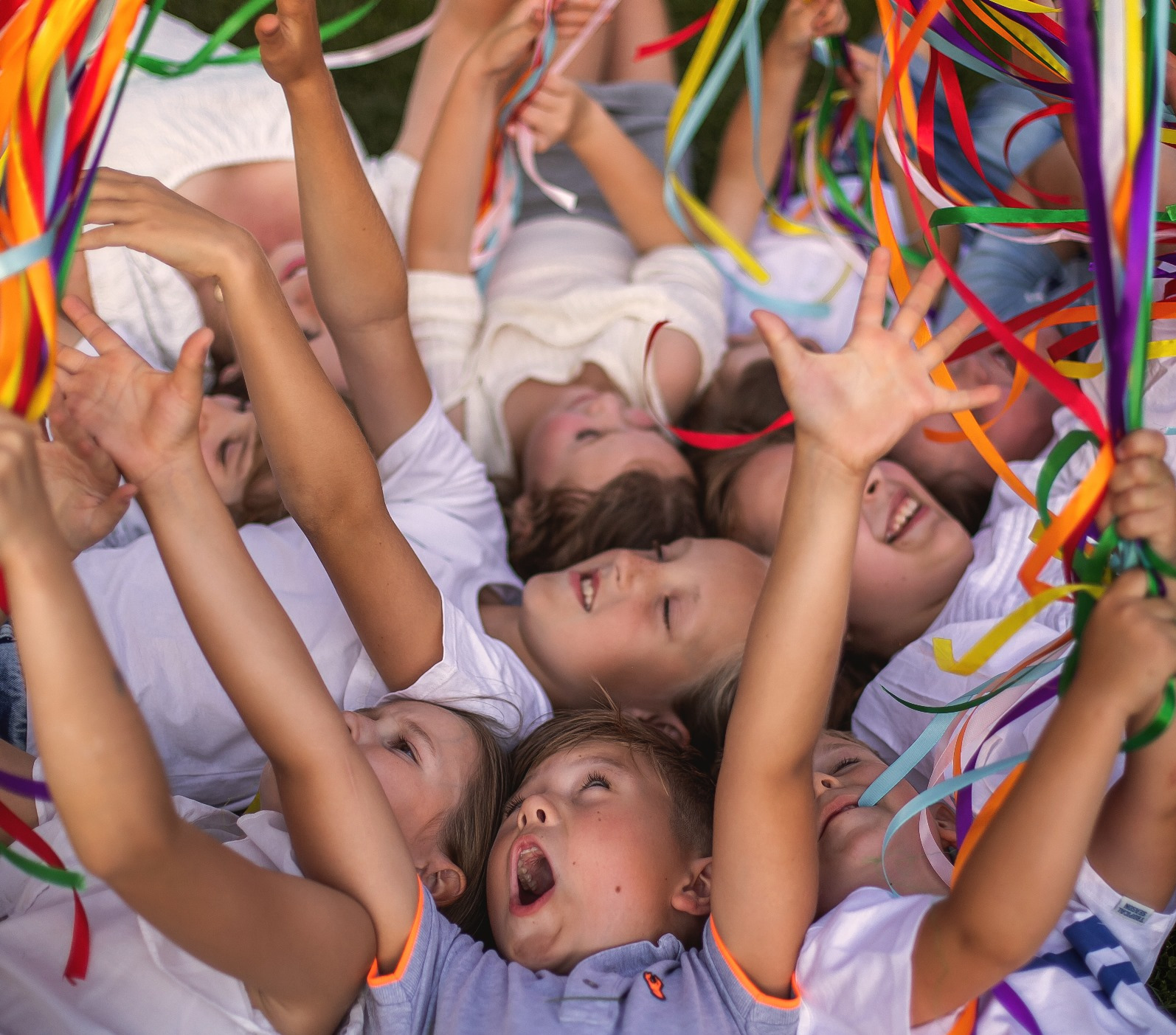 Grupa roześmianych dzieci leżących na trawie, trzymających kolorowe serpentyny, z otwartymi ustami, w jasnym, naturalnym świetle.
