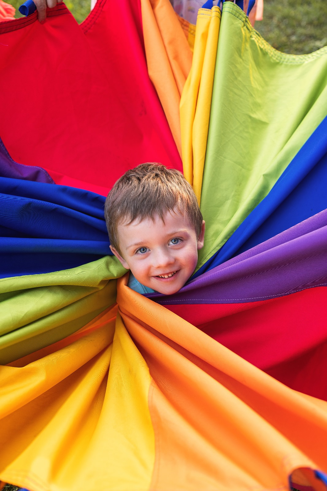 Uśmiechnięty chłopiec wystaje z kolorowej, tęczowej chusty animacyjnej, używanej podczas zabaw i gier na imprezach dla dzieci.