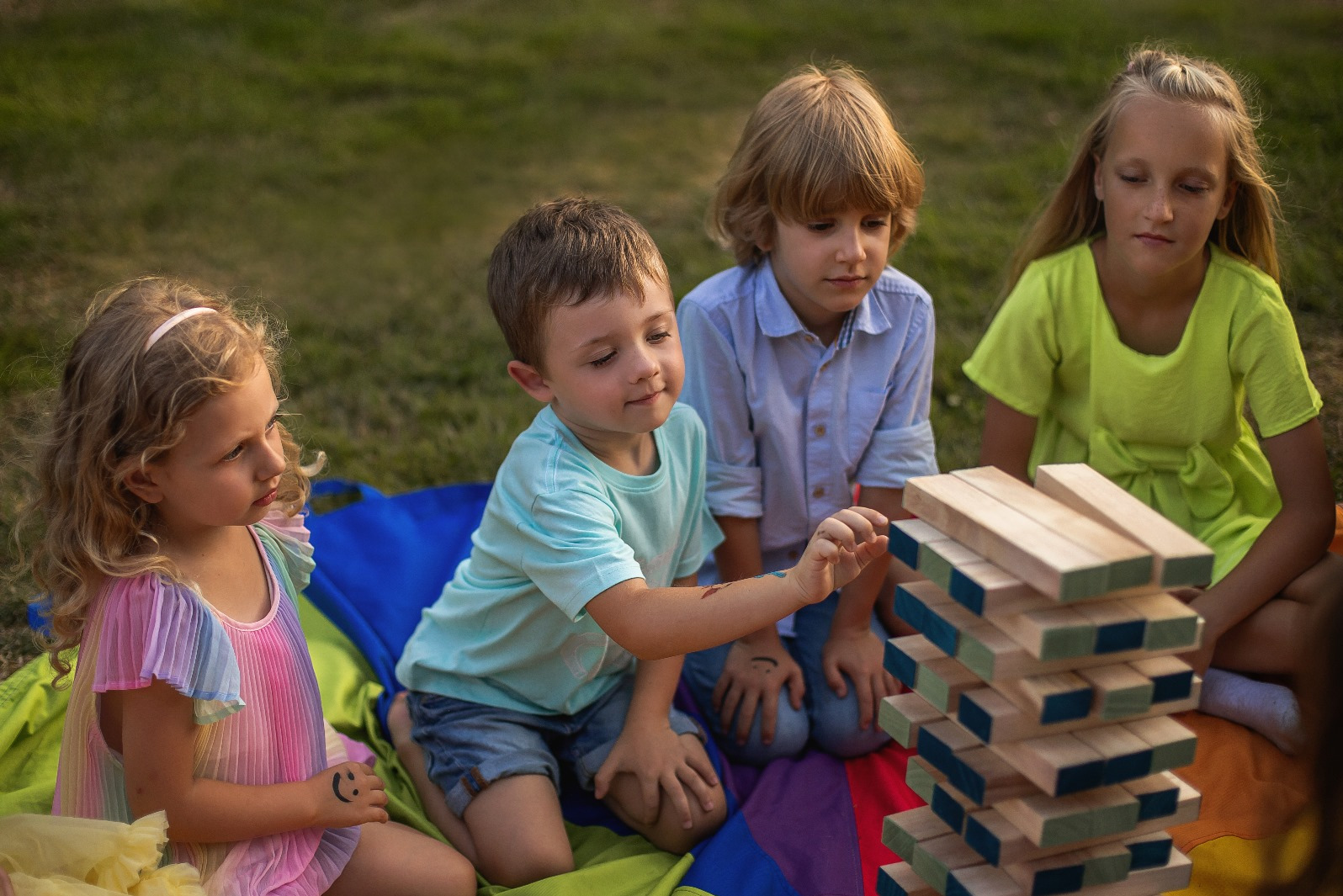 Czwórka dzieci siedzi na kocu grając w drewnianą wieżę Jenga na trawie w plenerze. Chłopiec w niebieskiej koszulce wyciąga klocek.