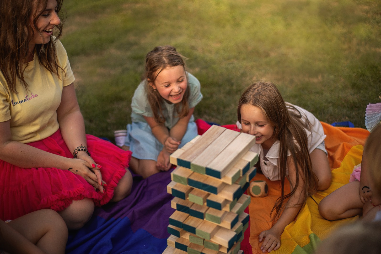 Radosne dzieci grają w drewnianą wieżę na kolorowym kocu, pod czujnym okiem animatorki w żółtej koszulce. Zabawa na trawie w słoneczny dzień.