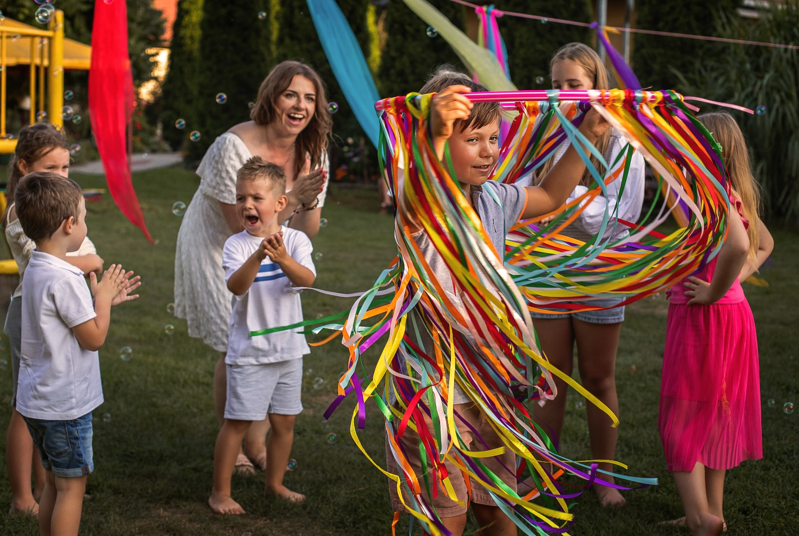 Radosne dzieci na zielonej trawie, w tle kobieta klaszcze. Chłopiec trzyma kolorową obręcz z wstążkami, a w powietrzu unoszą się bańki mydlane. Uśmiechy i dobra zabawa.