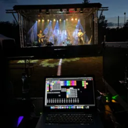 Zespół muzyczny na scenie oświetlonej reflektorami, widok zza laptopa z oprogramowaniem do sterowania oświetleniem, wieczorne ujęcie.