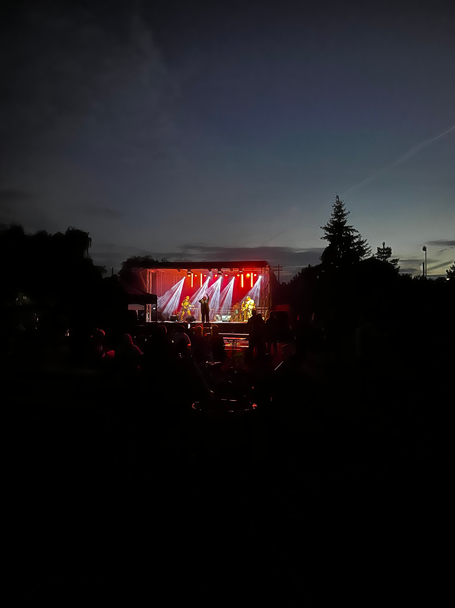 Zespół muzyczny na scenie oświetlonej czerwonymi reflektorami podczas wieczornego koncertu plenerowego, widoczni widzowie w tle.