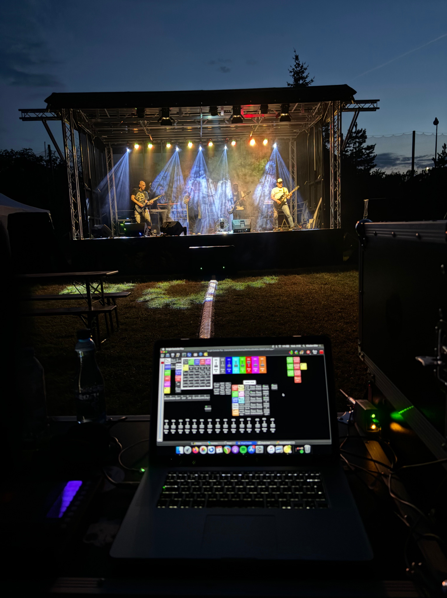 Zespół muzyczny na scenie oświetlonej reflektorami, widok zza laptopa z oprogramowaniem do sterowania oświetleniem, wieczorne ujęcie.