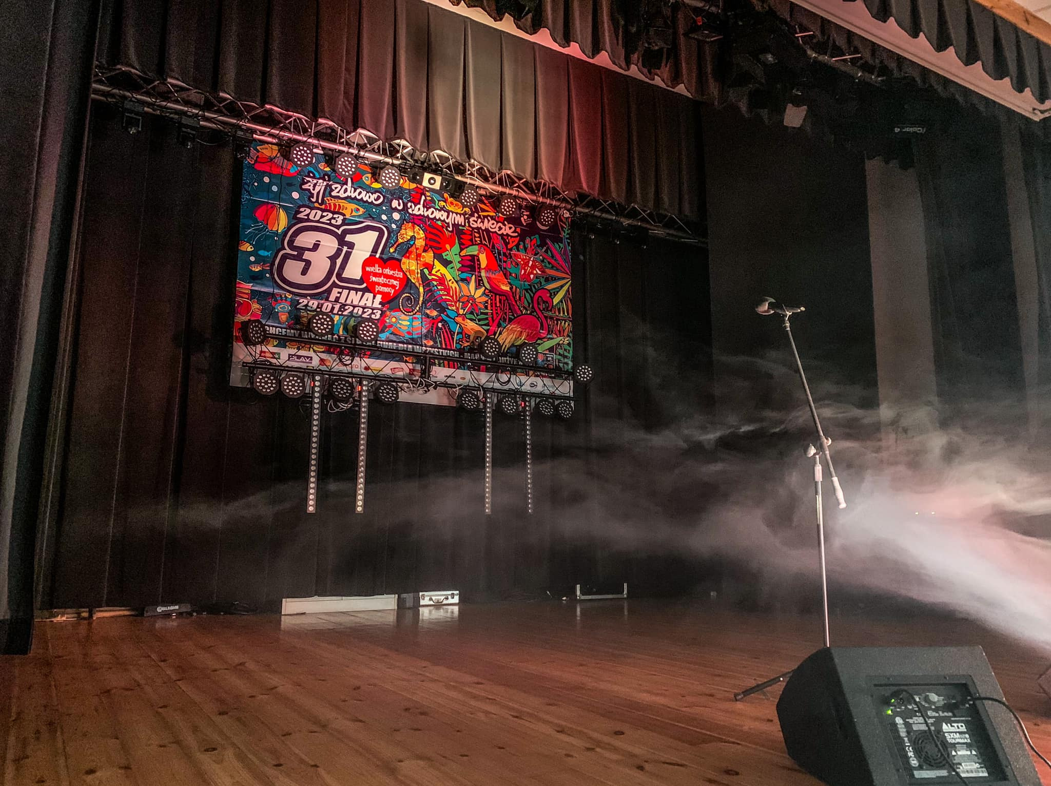 Scena przed wydarzeniem z plakatem WOŚP, oświetleniem i nagłośnieniem, unosząca się mgła sceniczna, mikrofon na statywie i głośnik na drewnianej podłodze.