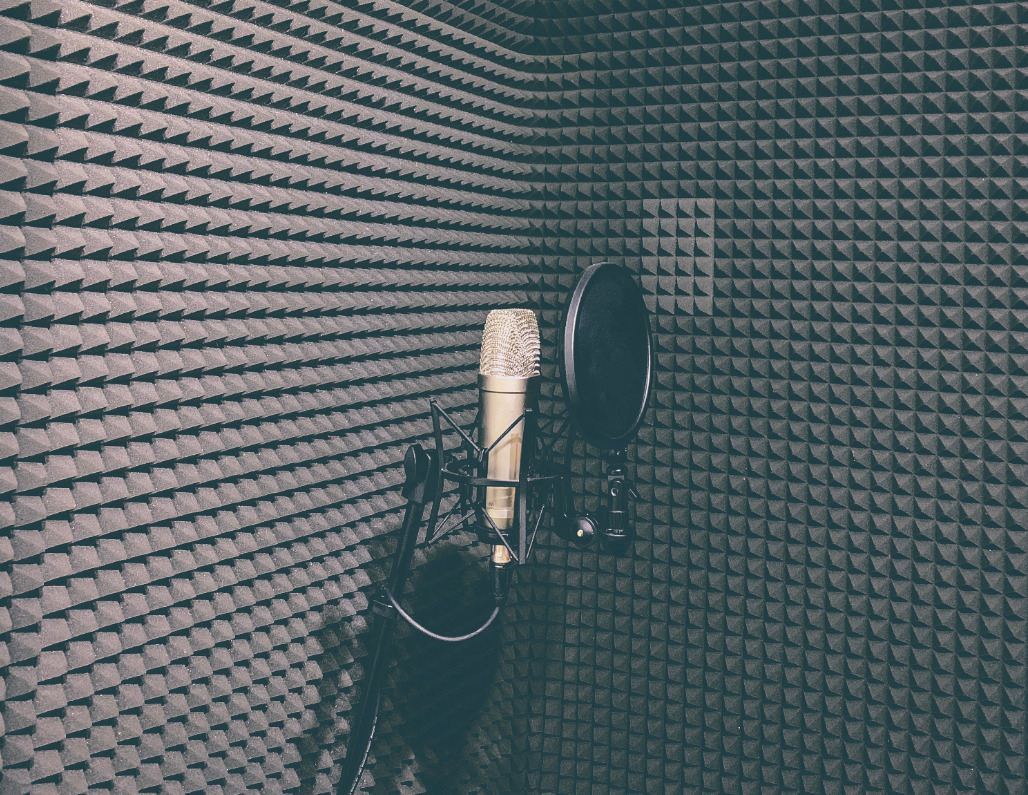 Złoty mikrofon studyjny z pop filtrem na tle ścian wyłożonych piramidkami z pianki akustycznej w rogu pomieszczenia.