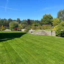 Zielona Zatoka Ewa Boniecka - Rozległy, zadbany trawnik z wyraźnymi pasami po koszeniu, prowadzący do kamiennego murku z roślinami i białej ławeczki w tle, pod błękitnym niebem.