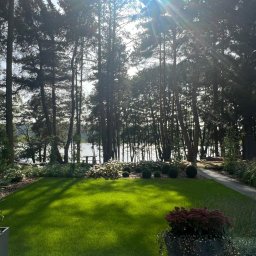 Zielona Zatoka Ewa Boniecka - Wygląd ogrodu z idealnie skoszoną trawą, kulistymi krzewami i widokiem na jezioro w tle, widziany z tarasu w słoneczny dzień.