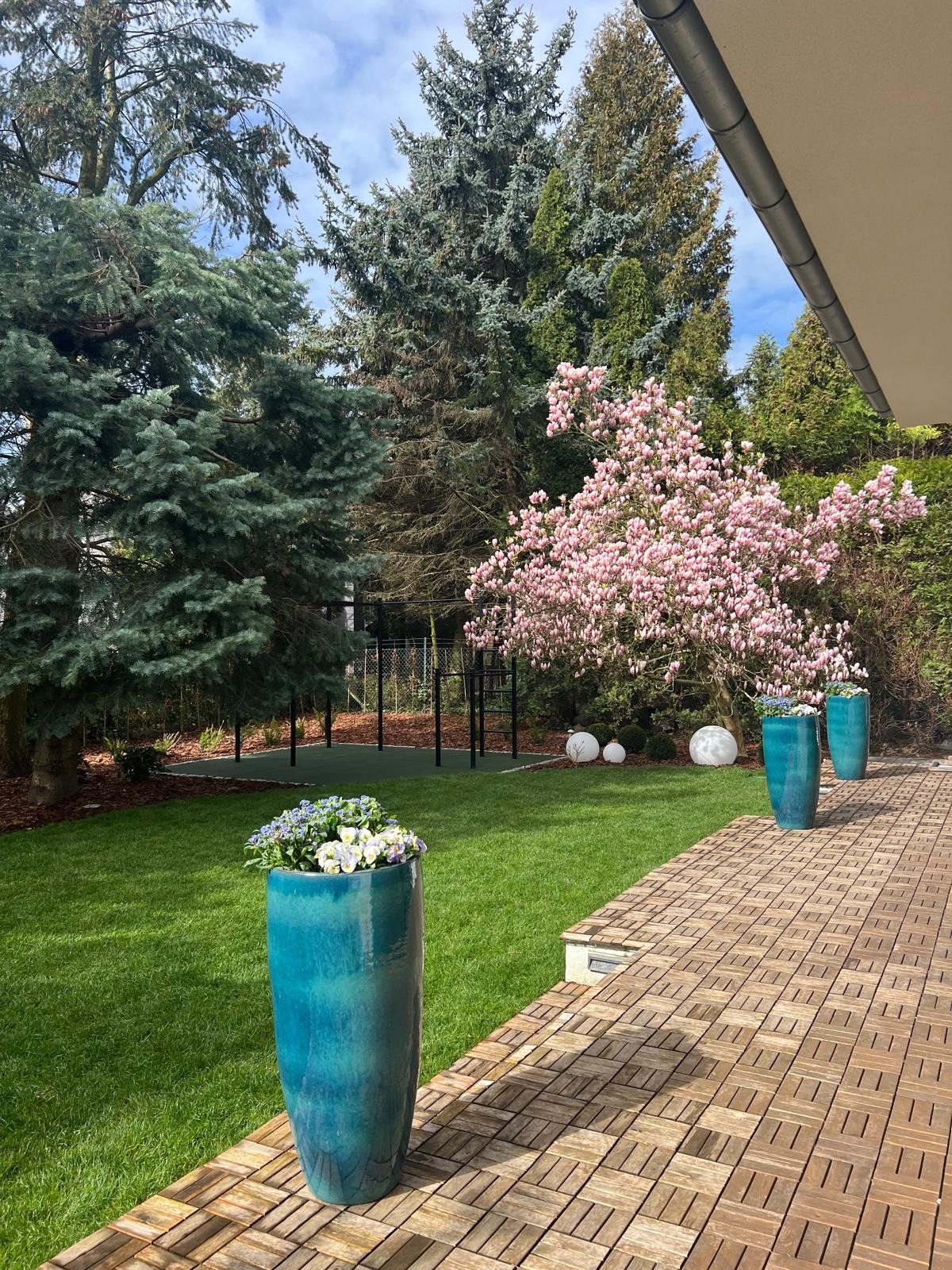 Utrzymany w zieleni ogród z kwitnącą magnolią, turkusowymi donicami z kwiatami i drewnianą kostką brukową na tarasie, w tle plac zabaw.
