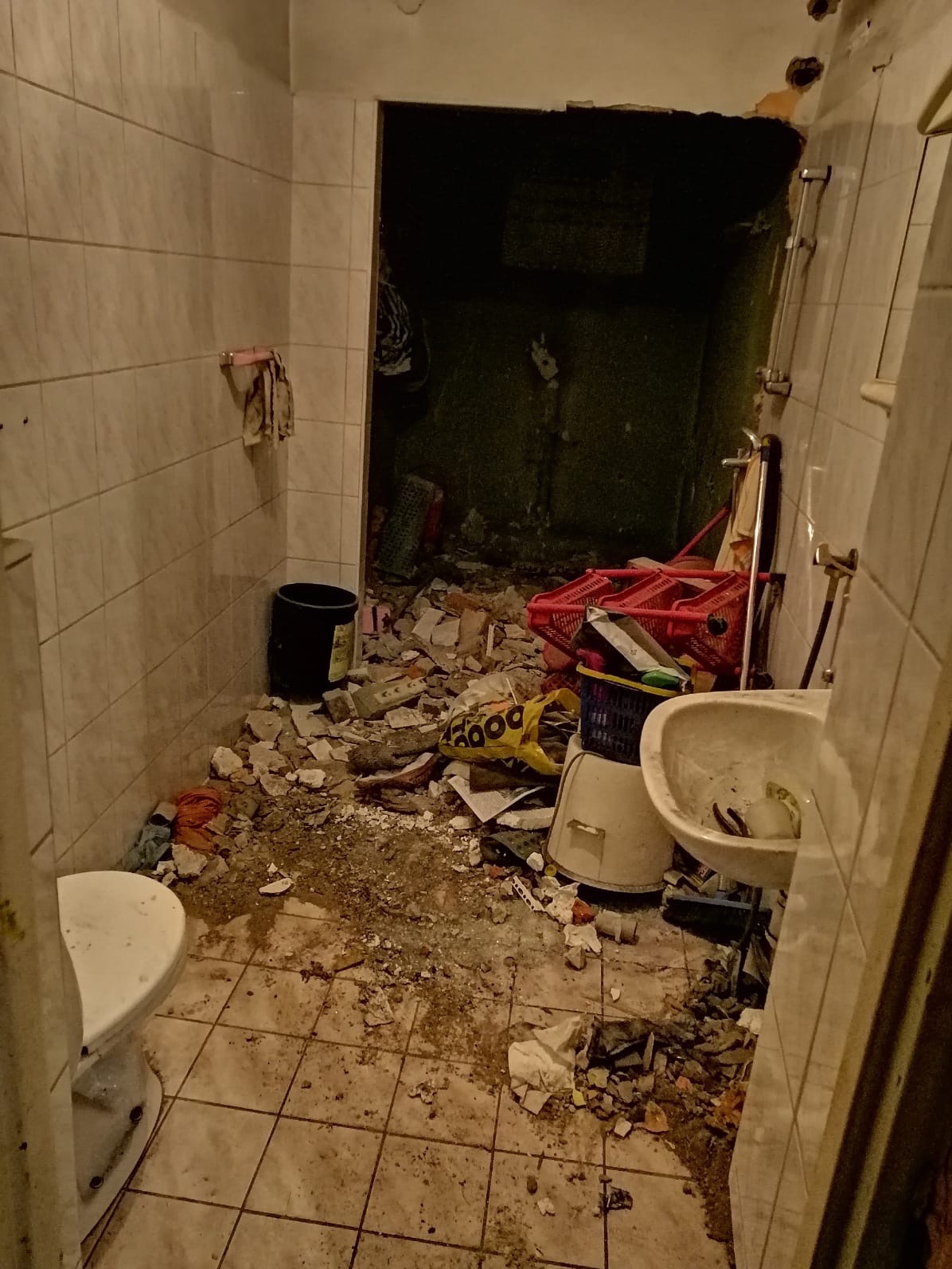 Zdemolowana łazienka z gruzem, resztkami wyposażenia, koszami i torbami na podłodze, widok na sąsiednie pomieszczenie bez drzwi, ślady demontażu na ścianach.