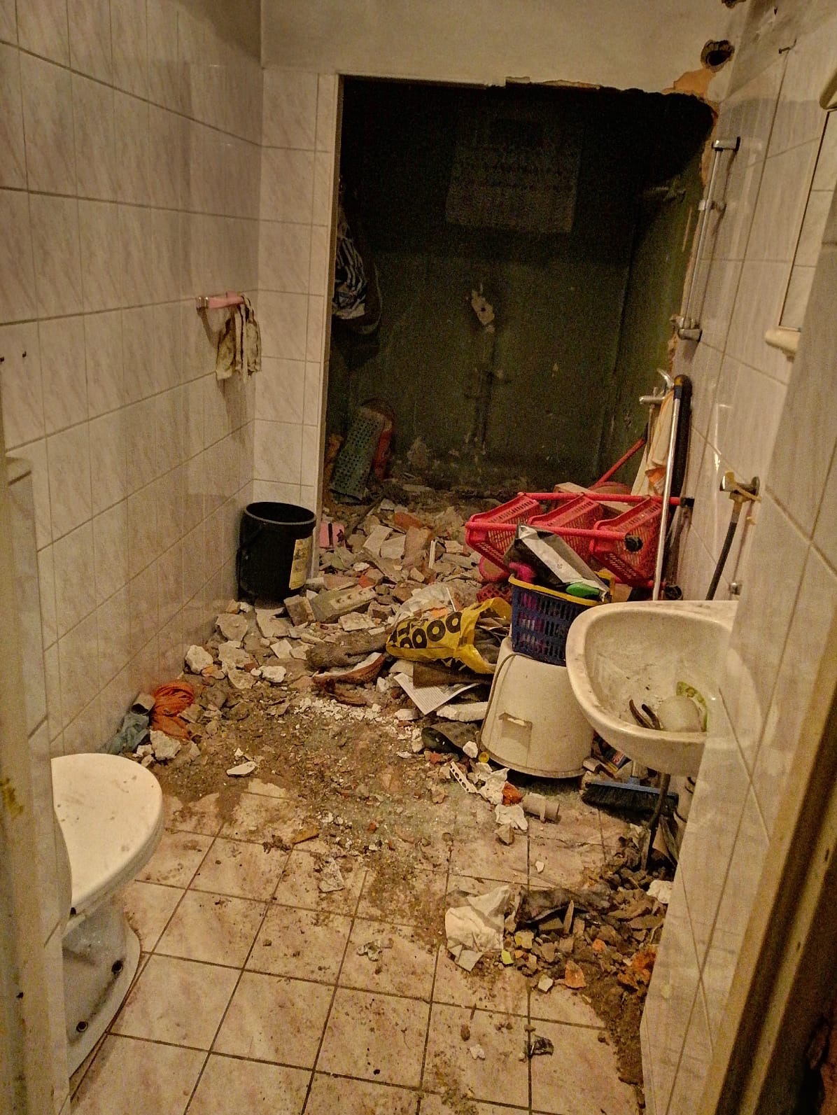 Zdemolowana łazienka w trakcie demontażu, widoczne resztki płytek, gruz, umywalka, toaleta, kosze i torba na śmieci, otwarte przejście do kolejnego, zagraconego pomieszczenia.