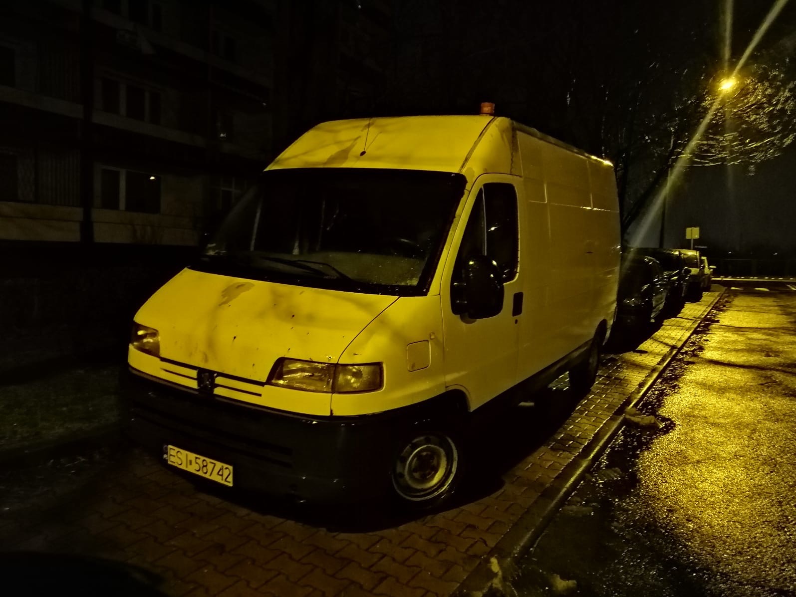 Żółty samochód dostawczy Peugeot zaparkowany nocą na mokrym chodniku, widoczne ślady zabrudzeń na karoserii i pomarańczowe światło ostrzegawcze na dachu.