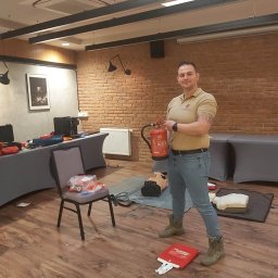 Bartosz Wiśniewski Usługi Medyczne WIS-MED - Instruktor z tatuażem na ramieniu, trzyma gaśnicę w sali szkoleniowej z manekinami do resuscytacji i apteczkami pierwszej pomocy.