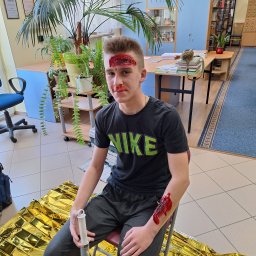 Bartosz Wiśniewski Usługi Medyczne WIS-MED - Młody mężczyzna z realistyczną symulacją ran na czole i ręce, siedzący na krześle na tle sali z roślinami i regałami, obok leży koc termiczny i strzykawka bez igły, prawdopodobnie w trakcie...