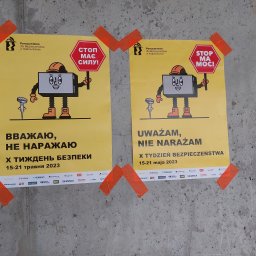 Bartosz Wiśniewski Usługi Medyczne WIS-MED - Dwa plakaty informacyjne z kreskówkowymi postaciami w kaskach, naklejone pomarańczową taśmą na szarym, surowym betonie. Plakaty promują tydzień bezpieczeństwa pracy w budownictwie, z hasłami...