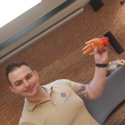 Bartosz Wiśniewski Usługi Medyczne WIS-MED - Instruktor z uśmiechem prezentuje pomarańczową atrapę dłoni z symulacją urazu podczas kursu pierwszej pomocy, widoczne logo WIS-MED na koszulce.