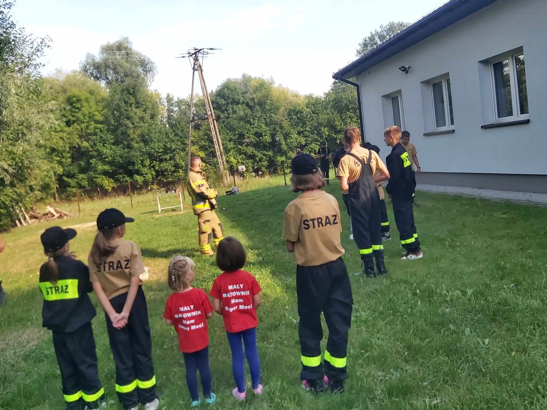 Grupa dzieci i dorosłych w strojach strażackich na trawniku przed budynkiem, prawdopodobnie podczas szkolenia lub pokazu.