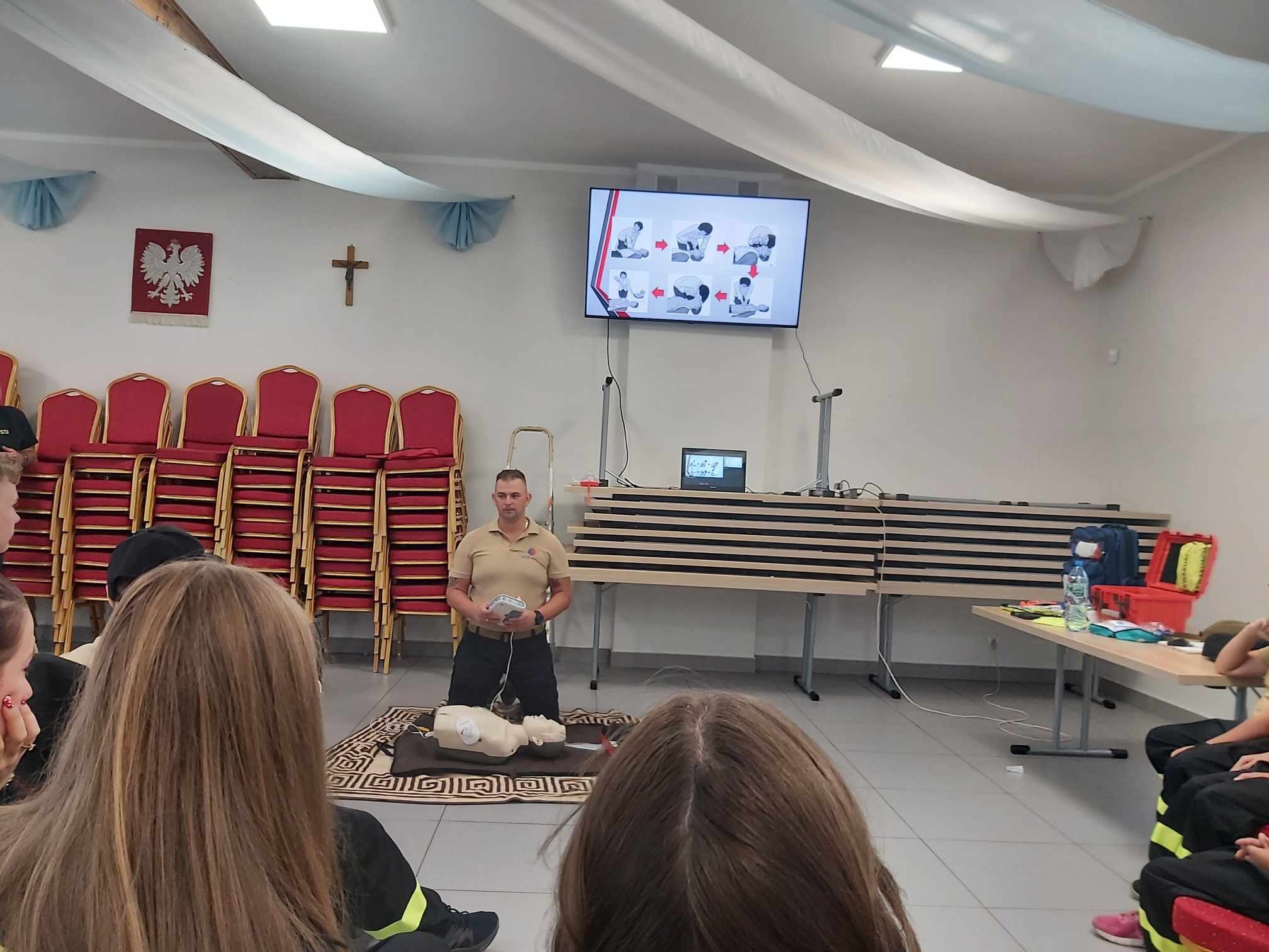 Instruktor demonstruje użycie defibrylatora na manekinie podczas zajęć z pierwszej pomocy, uczestnicy siedzą na podłodze, na ścianie wisi ekran z instrukcjami. W tle godło Polski i krzyż.