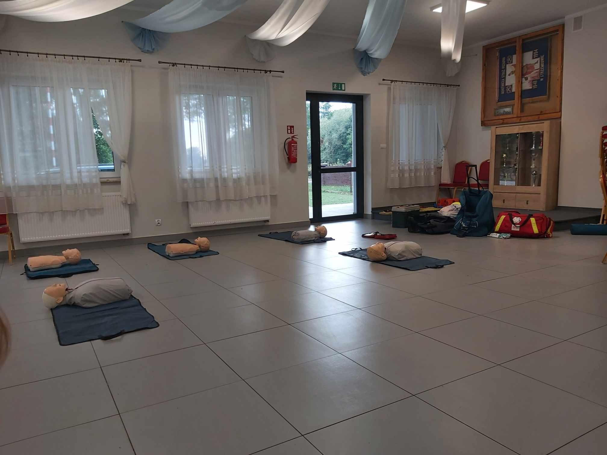 Sala z manekinami na matach, przygotowana do szkolenia z pierwszej pomocy. Widoczny gaśnica, apteczka i torby ratunkowe.