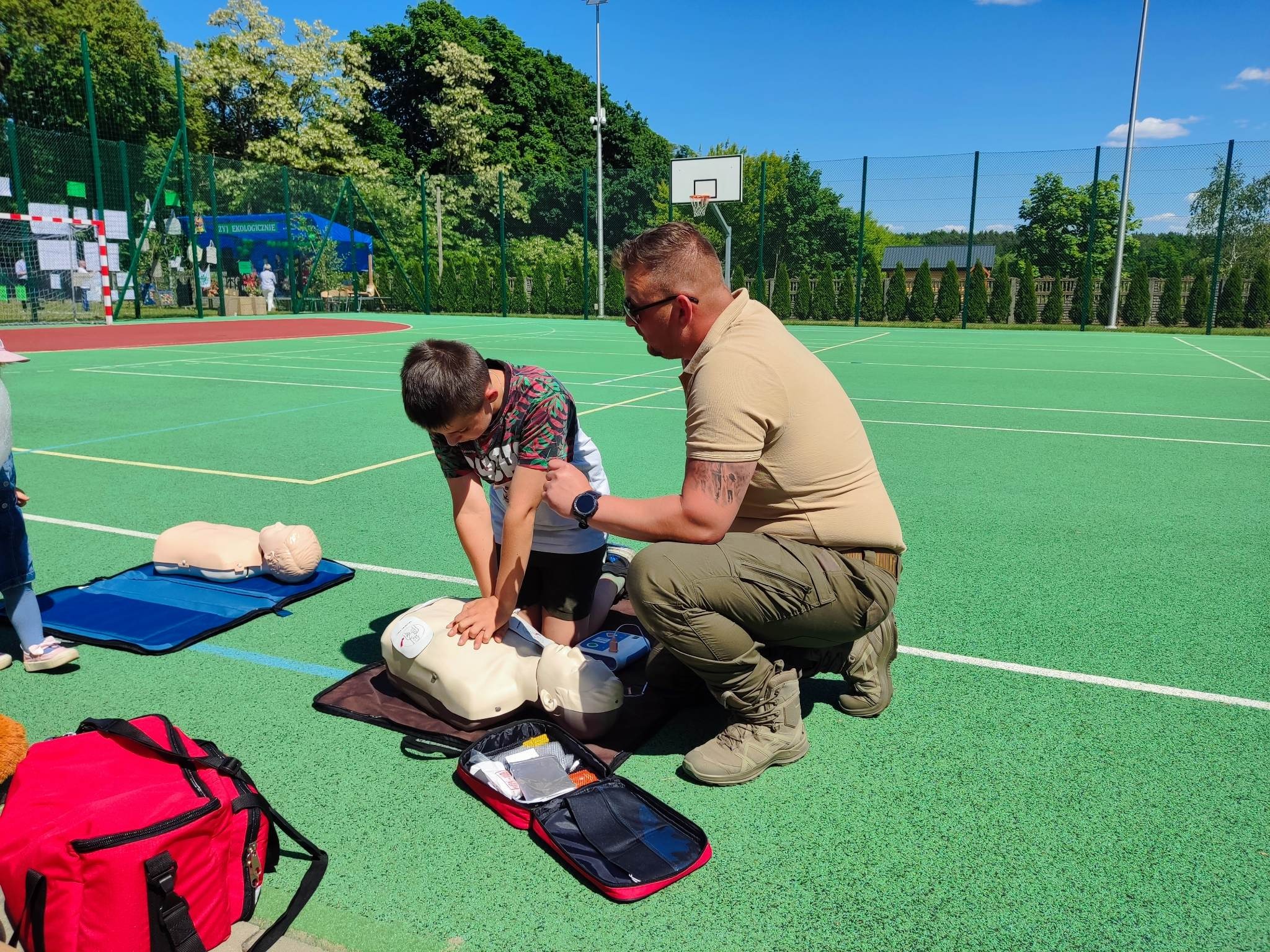 Chłopiec ćwiczy resuscytację krążeniowo-oddechową na manekinie pod nadzorem instruktora na zielonym boisku sportowym, obok torba z zestawem pierwszej pomocy.