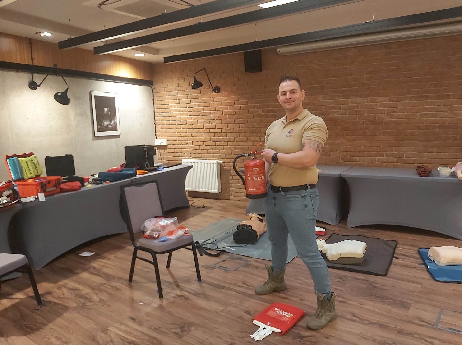 Instruktor z tatuażem na ramieniu, trzyma gaśnicę w sali szkoleniowej z manekinami do resuscytacji i apteczkami pierwszej pomocy.