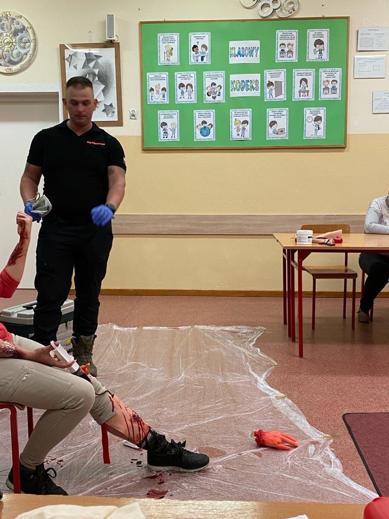 Instruktor w czarnym ubraniu i niebieskich rękawiczkach demonstruje opatrywanie krwawiącej rany na manekinie podczas symulacji w sali szkoleniowej. Na podłodze folia ochronna i odcięta sztuczna dłoń.
