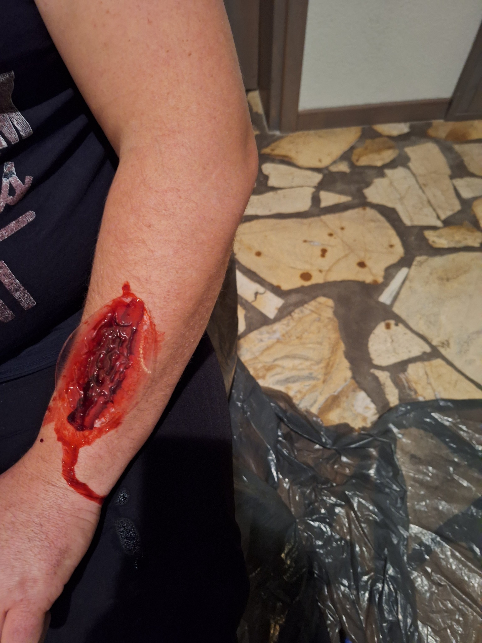 Ramię z realistyczną raną otwartą, krew spływa po skórze, widoczne elementy imitujące tkankę, tło z mozaiką podłogową i czarną folią.