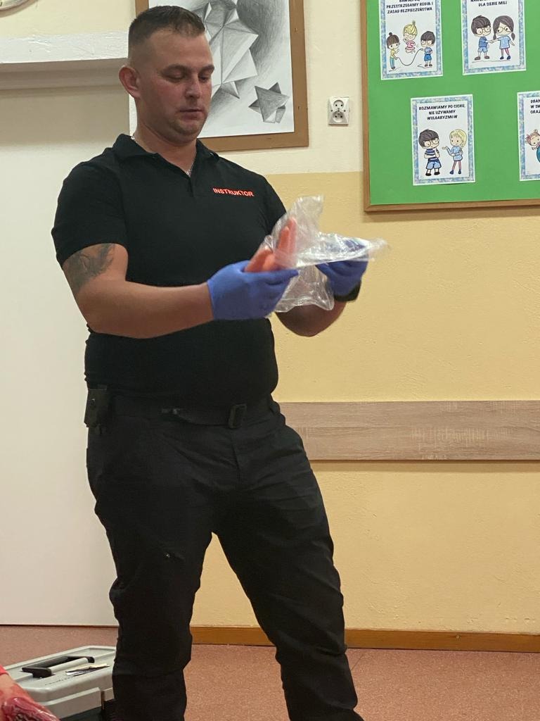 Instruktor w czarnej koszulce z napisem 'INSTRUKTOR' prezentuje manekin do nauki pierwszej pomocy, w rękawiczkach, na tle ściany z rysunkami edukacyjnymi.