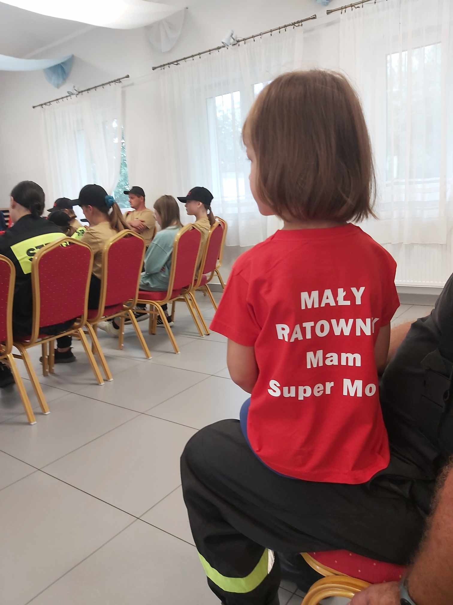 Dziewczynka w czerwonej koszulce z napisem 'Mały Ratownik, Mam Super Moc' siedzi na kolanach osoby w stroju strażackim, w tle grupa osób na krzesłach podczas szkolenia.