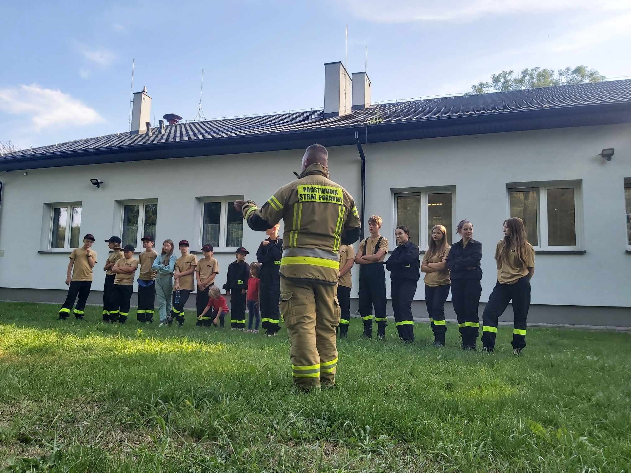 Strażak Państwowej Straży Pożarnej prowadzi szkolenie dla grupy młodzieży na trawniku przed budynkiem straży pożarnej.
