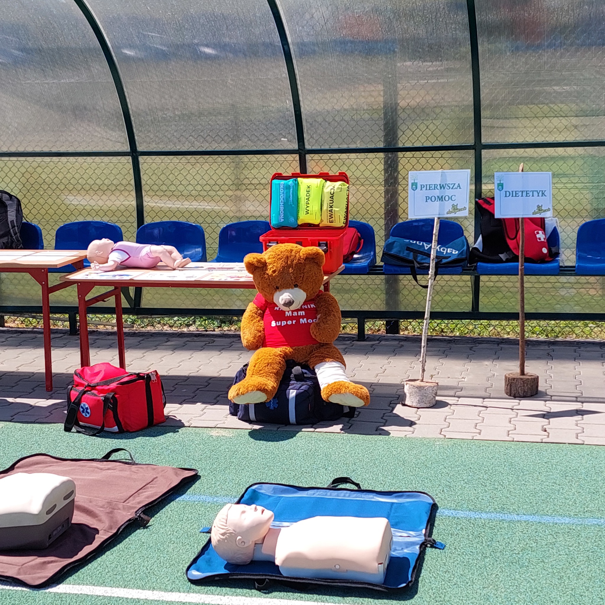 Scena demonstracyjna z manekinami do nauki pierwszej pomocy, pluszowy miś z zabandażowaną nogą siedzący na torbie, apteczka pierwszej pomocy i tabliczki informacyjne w tle, ustawione na sztucznej...