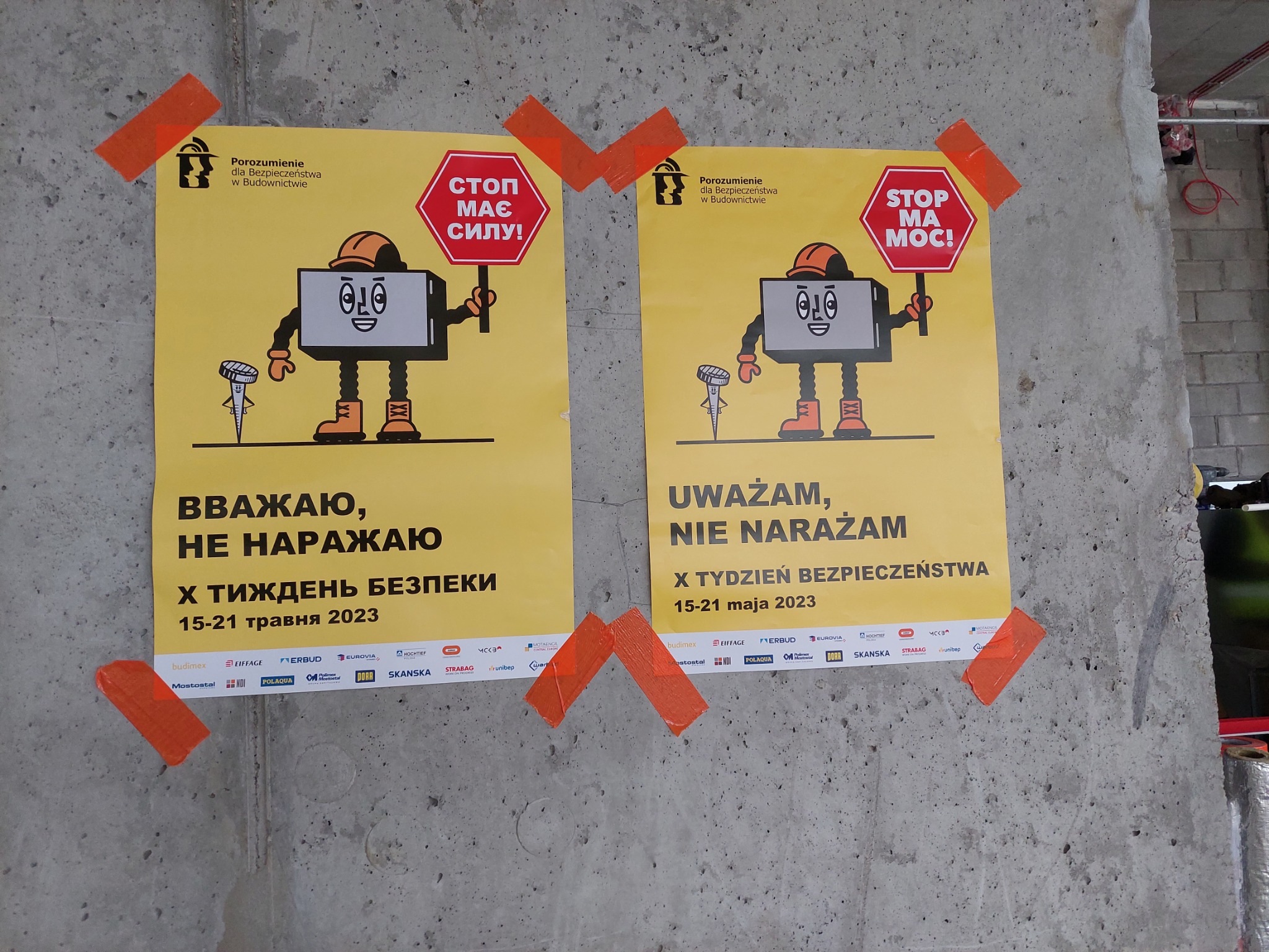 Dwa plakaty informacyjne z kreskówkowymi postaciami w kaskach, naklejone pomarańczową taśmą na szarym, surowym betonie. Plakaty promują tydzień bezpieczeństwa pracy w budownictwie, z hasłami...