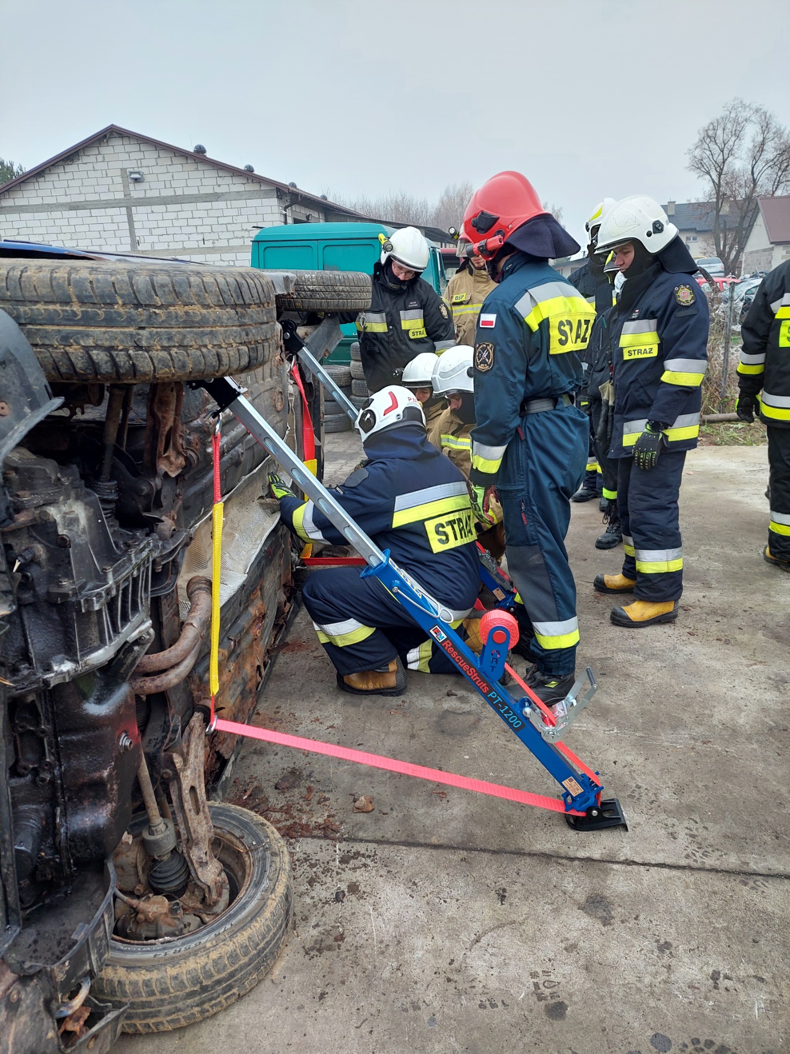 Strażacy w kombinezonach interweniują przy przewróconym samochodzie używając specjalistycznego sprzętu do stabilizacji pojazdu, widoczne pasy zabezpieczające i hydrauliczne rozpieracze.
