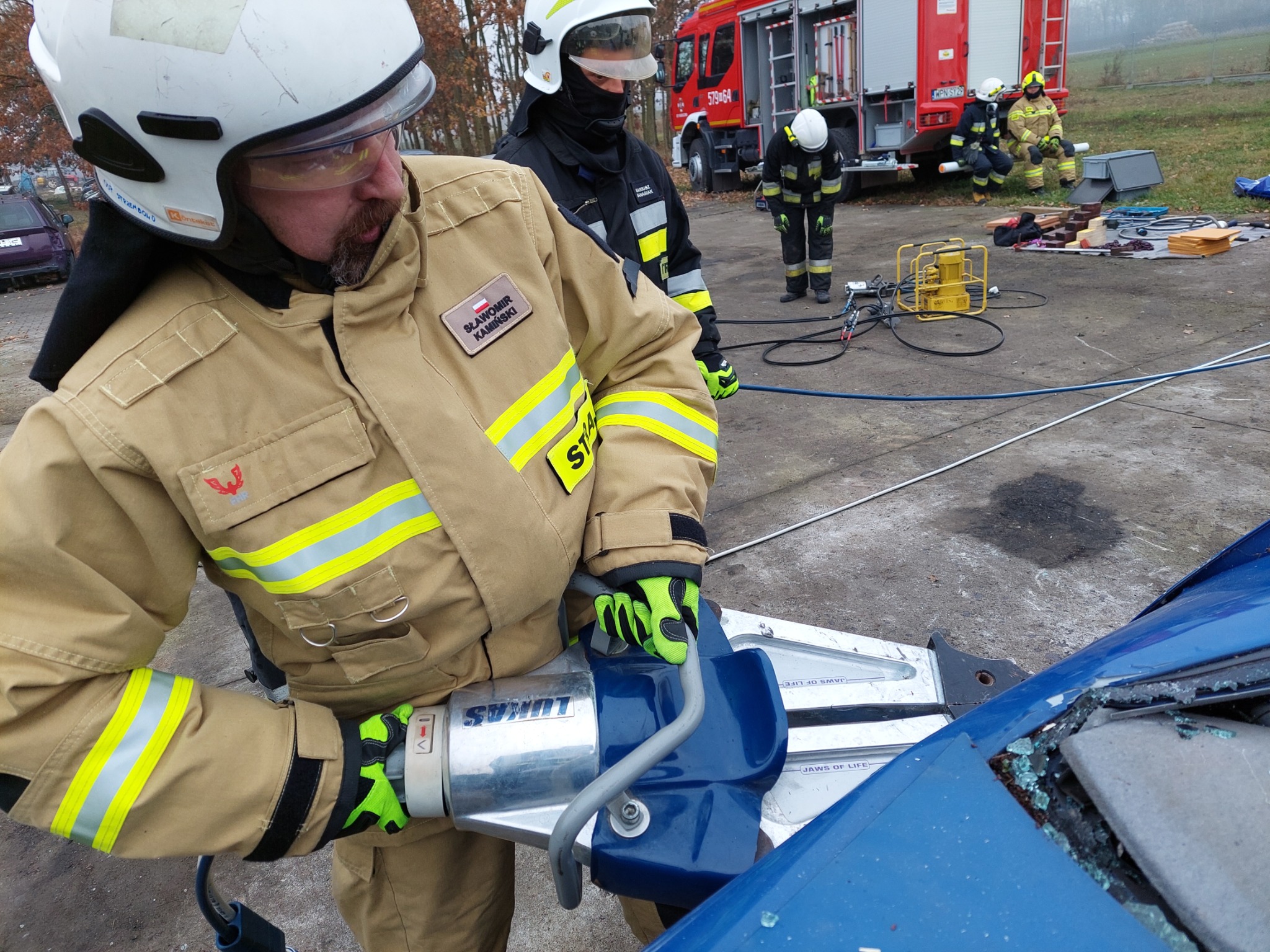 Strażak w umundurowaniu, z naszywką 'Sławomir Kamiński', używa hydraulicznych nożyc LUKAS do rozcinania wraku niebieskiego samochodu, w tle widać wóz strażacki i innych strażaków.