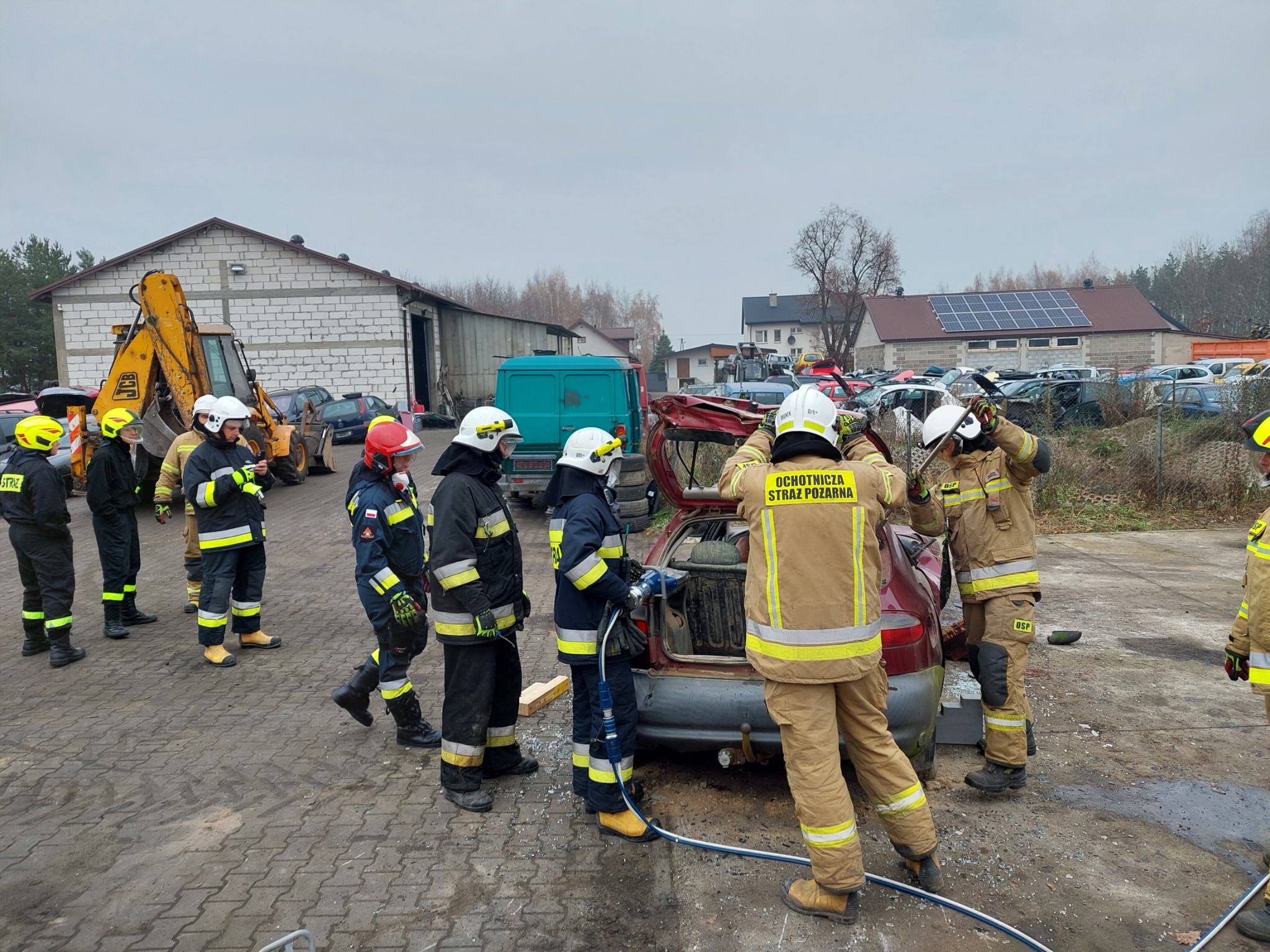 Grupa strażaków ochotników w umundurowaniu ćwiczy przy rozbitym samochodzie na placu manewrowym, w tle budynki i zaparkowane pojazdy.