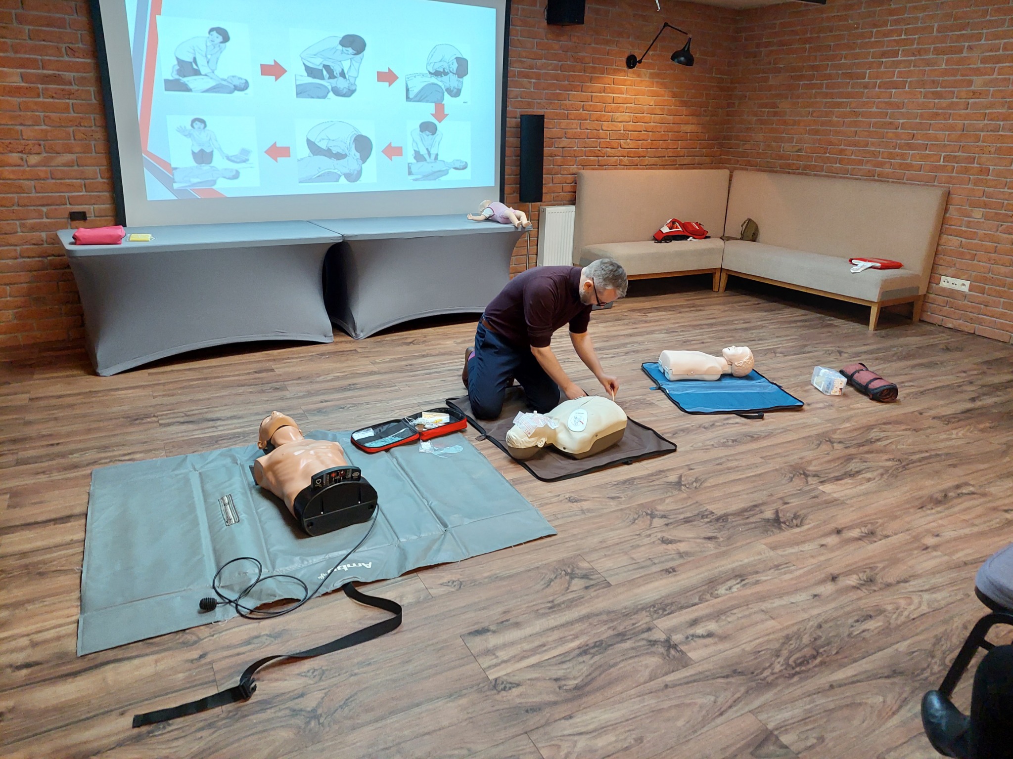 Instruktor w maseczce demonstruje resuscytację na manekinie podczas zajęć, na ścianie rzutnik wyświetla schemat postępowania, w tle ceglana ściana i beżowa sofa.