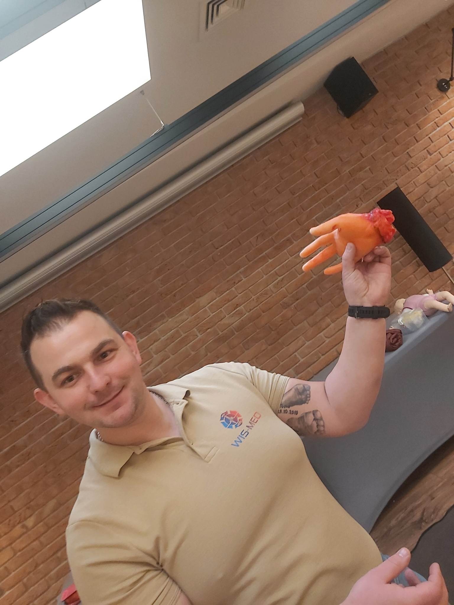 Instruktor z uśmiechem prezentuje pomarańczową atrapę dłoni z symulacją urazu podczas kursu pierwszej pomocy, widoczne logo WIS-MED na koszulce.
