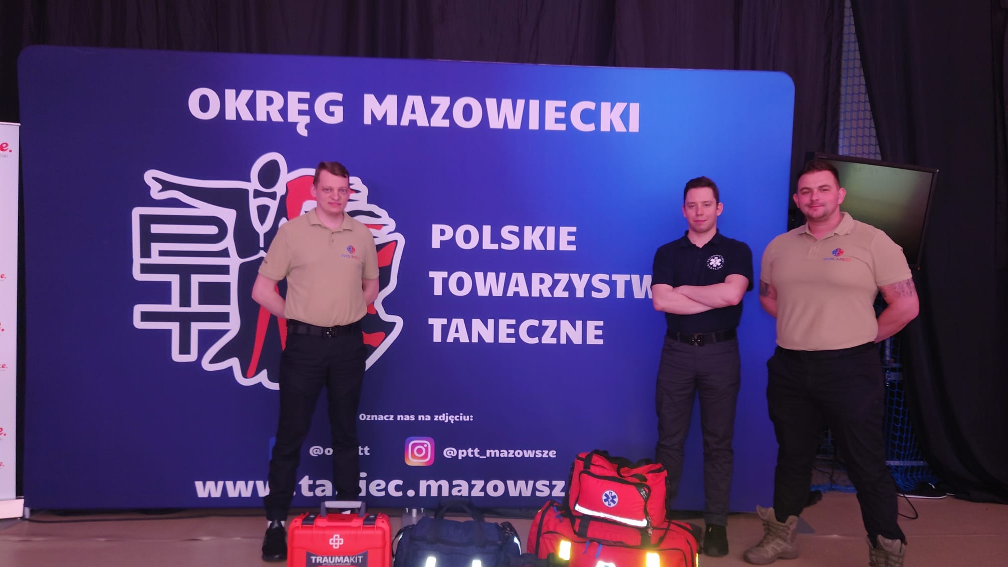 Trzech mężczyzn w uniformach WISS MED stoi na tle baneru 'Okręg Mazowiecki Polskiego Towarzystwa Tanecznego', z torbami medycznymi i zestawem TRAUMAKIT na podłodze.