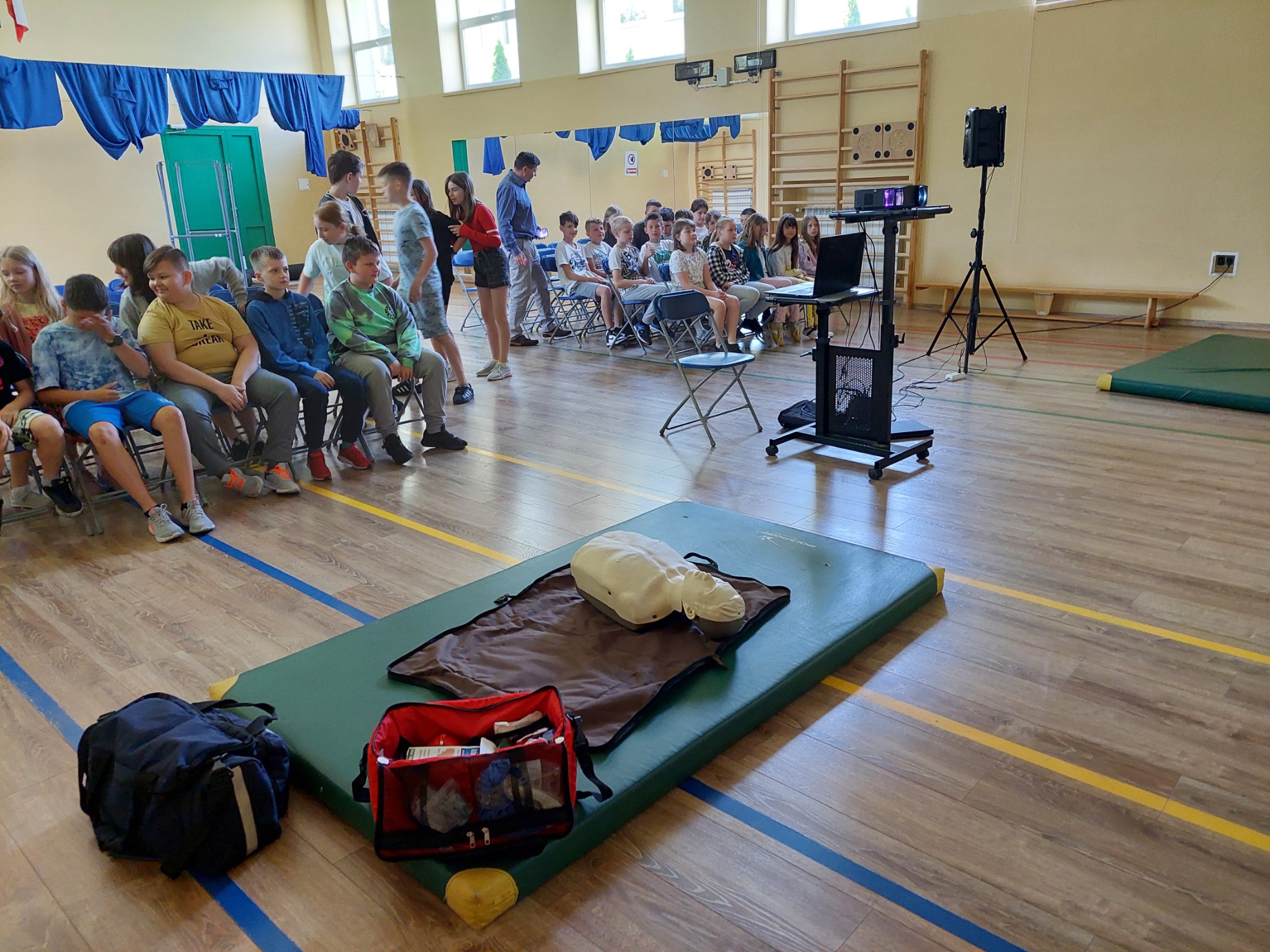 Grupa uczniów uczestniczy w szkoleniu z pierwszej pomocy w sali gimnastycznej, z manekinem do resuscytacji i torbą medyczną na macie, projektor i laptop na stoliku z boku.