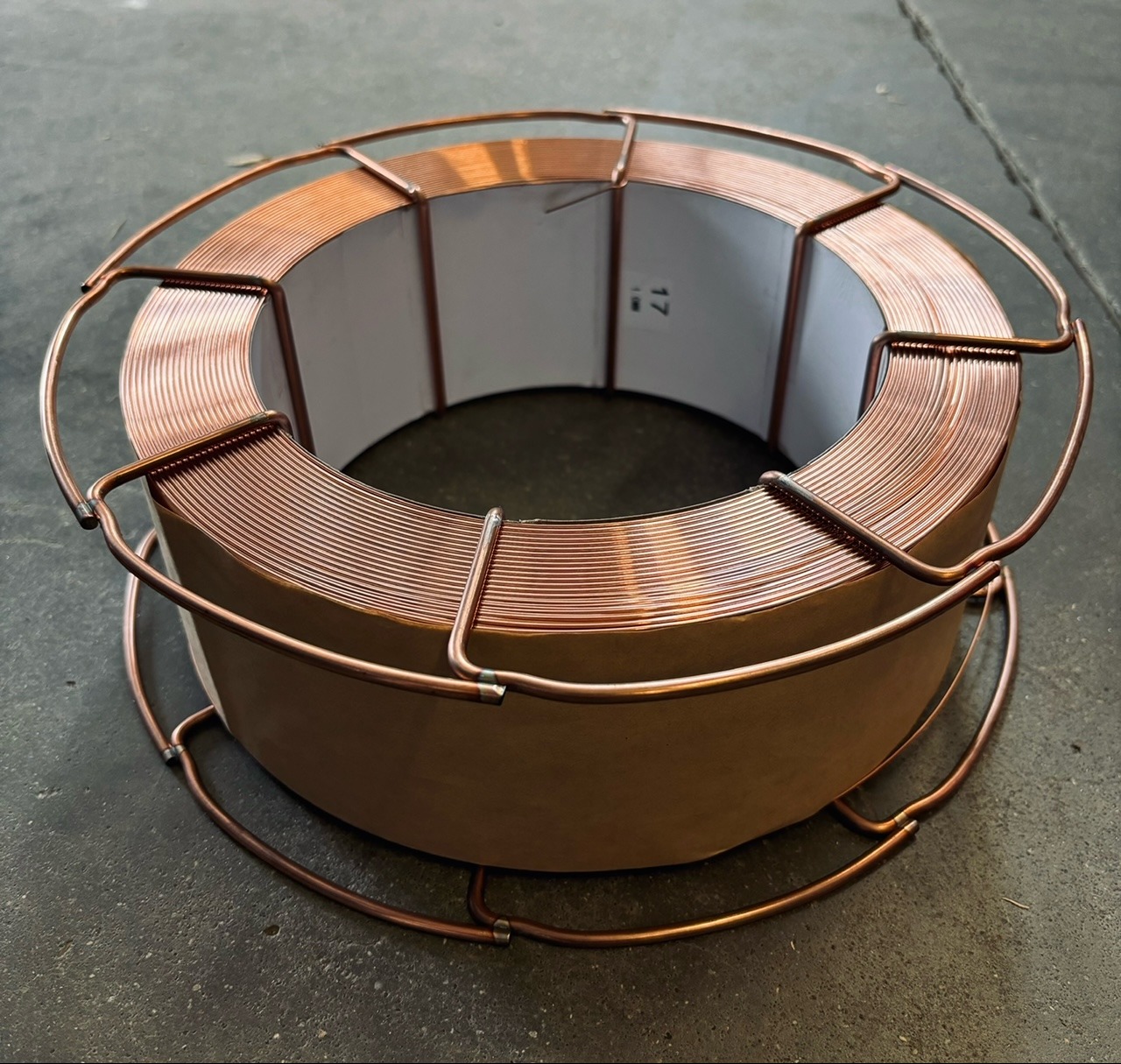 Szpula miedzianego drutu spawalniczego w metalowym koszu ochronnym, widoczna numeracja na kartonowej osłonie drutu.