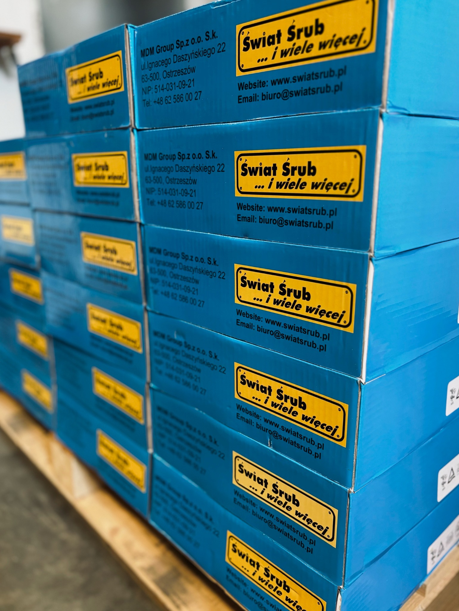 Stos niebieskich pudełek z logo 'Świat Śrub ... i wiele więcej' na drewnianej palecie, widoczne dane kontaktowe firmy.