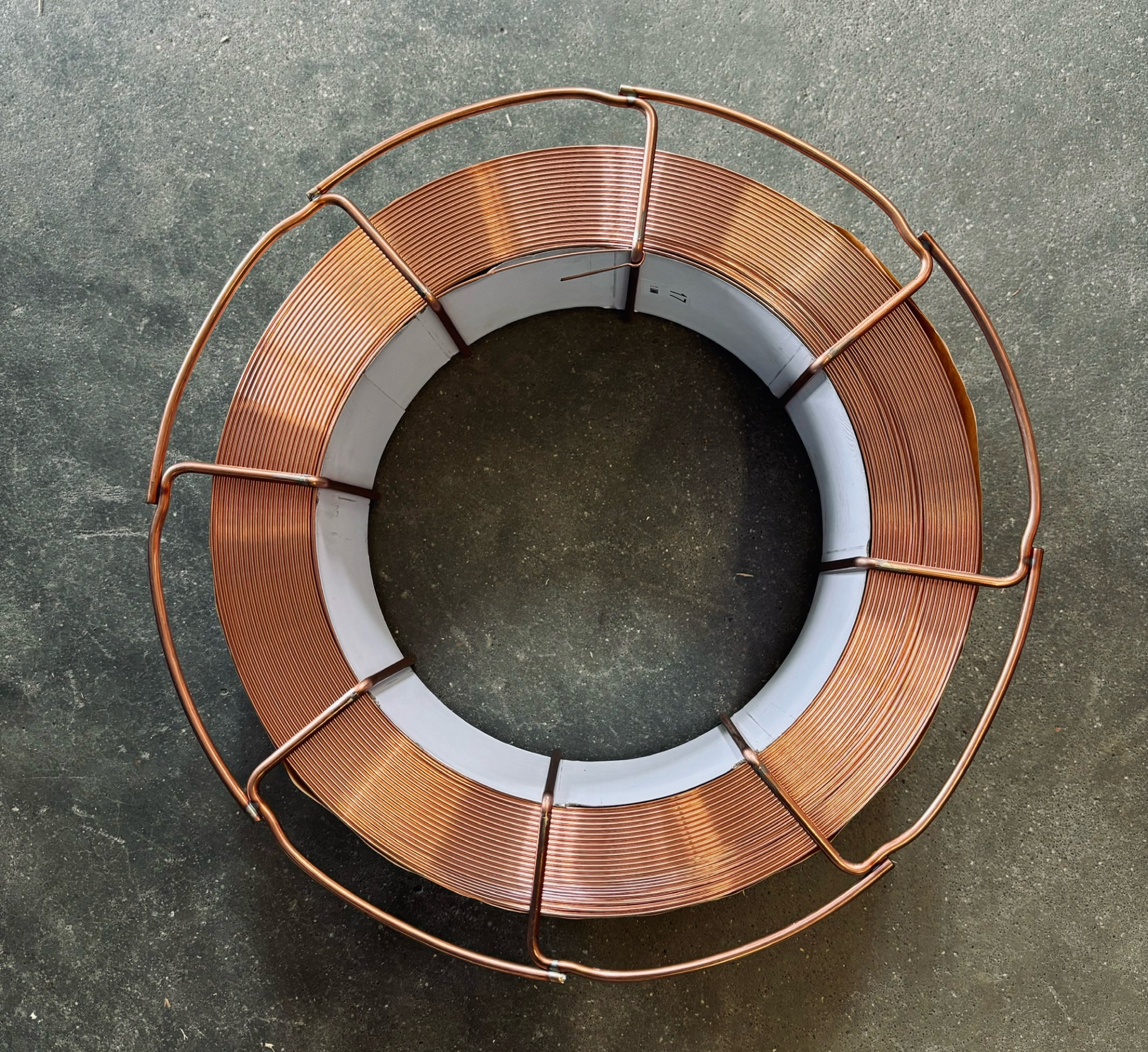 Szpula z grubym, miedzianym drutem spawalniczym, umieszczona w metalowym koszu ochronnym, na szarym, betonowym tle.