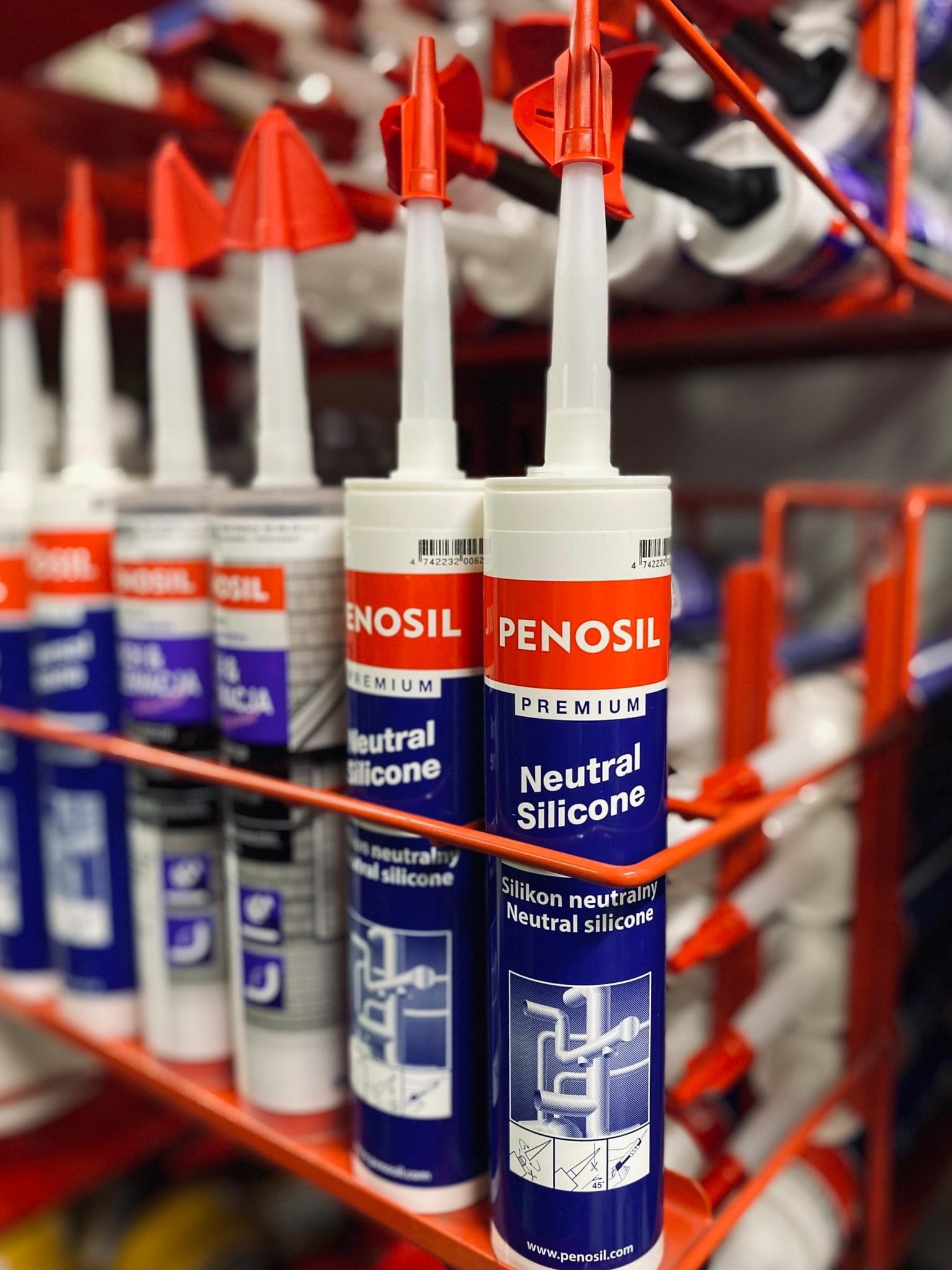 Rząd tub z neutralnym silikonem Penosil Premium na czerwonym metalowym regale w sklepie, widoczne etykiety z instrukcjami i logo.