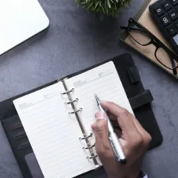 Osoba pisze srebrnym długopisem w otwartym, czarnym organizerze na tle biurka z laptopem, kalkulatorem, okularami i dekoracyjną rośliną.
