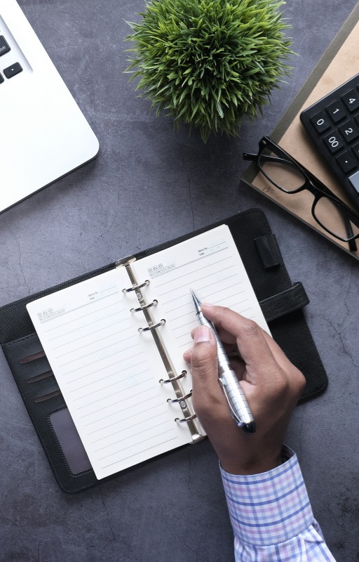 Osoba pisze srebrnym długopisem w otwartym, czarnym organizerze na tle biurka z laptopem, kalkulatorem, okularami i dekoracyjną rośliną.