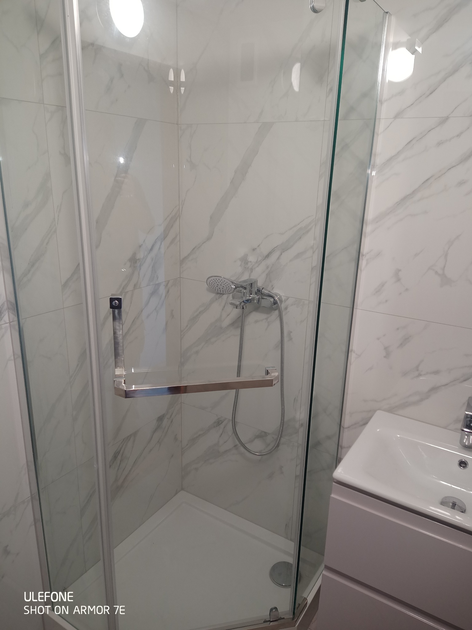 Kabina prysznicowa z chromowanym uchwytem i deszczownicą na tle białych marmurowych płytek w łazience z umywalką.