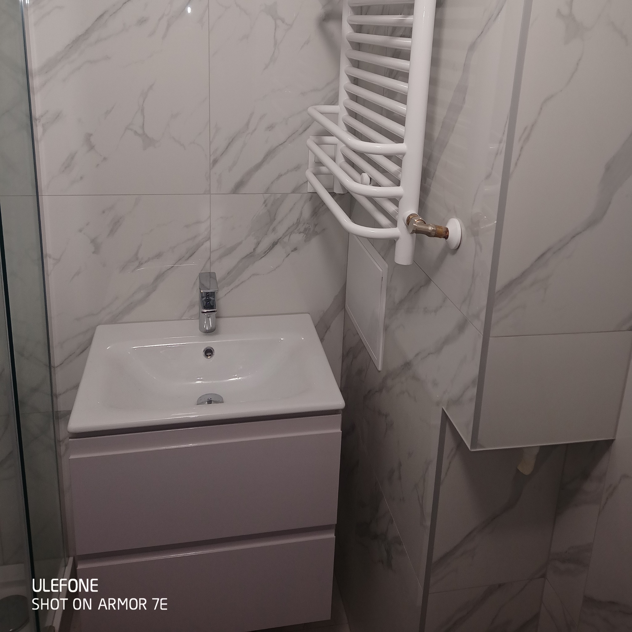 Nowoczesna łazienka z umywalką nablatową w szafce z szufladami, grzejnikiem drabinkowym i marmurowymi płytkami na ścianie.