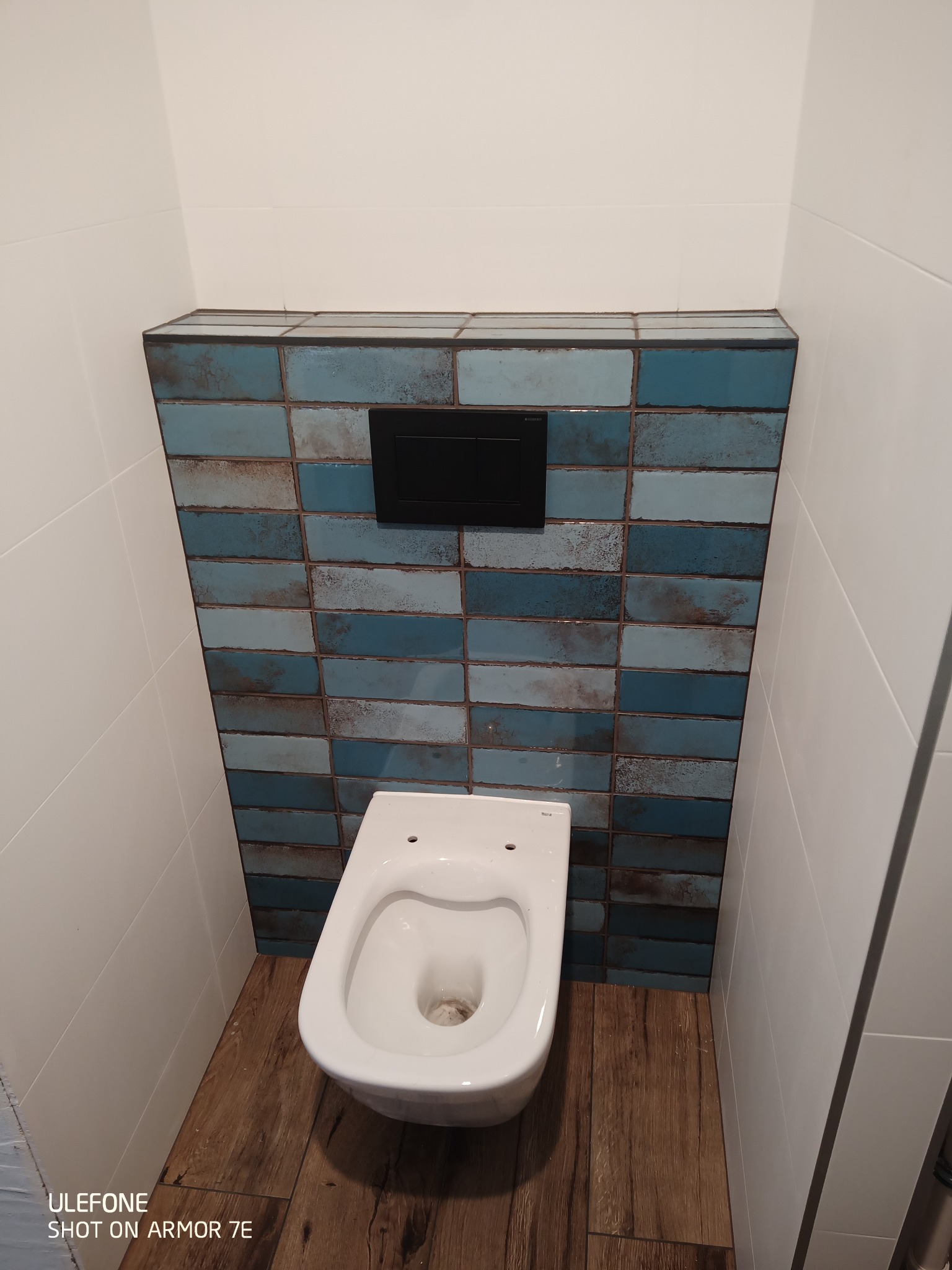 Wisząca toaleta z czarnym przyciskiem spłukującym, obudowana niebieskimi, rustykalnymi płytkami, widoczne drewnopodobne panele na podłodze i białe płytki na ścianach.