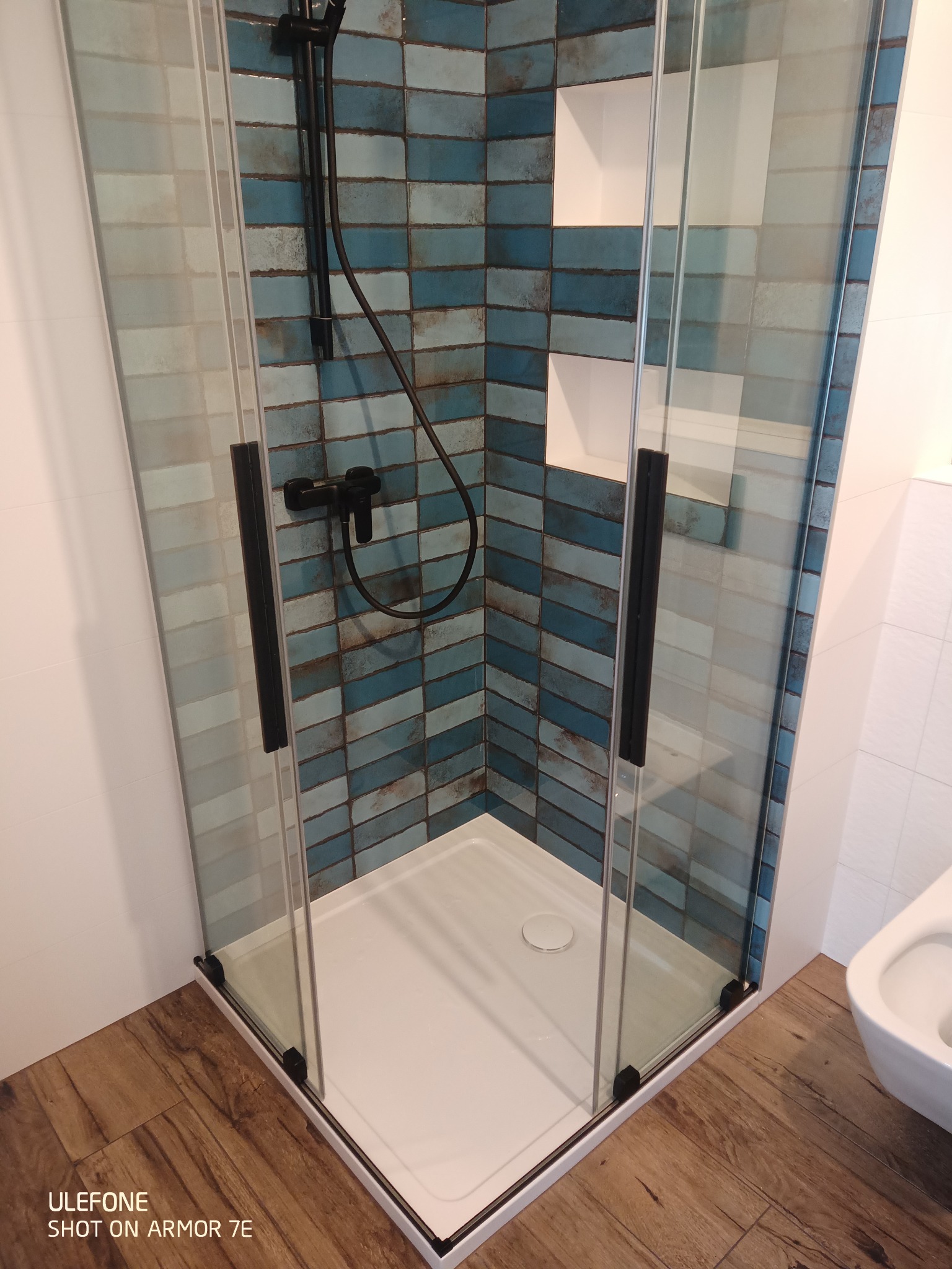 Kabina prysznicowa z niebieskimi płytkami ułożonymi w jodełkę, czarna armatura, biały brodzik i drewnopodobna podłoga.