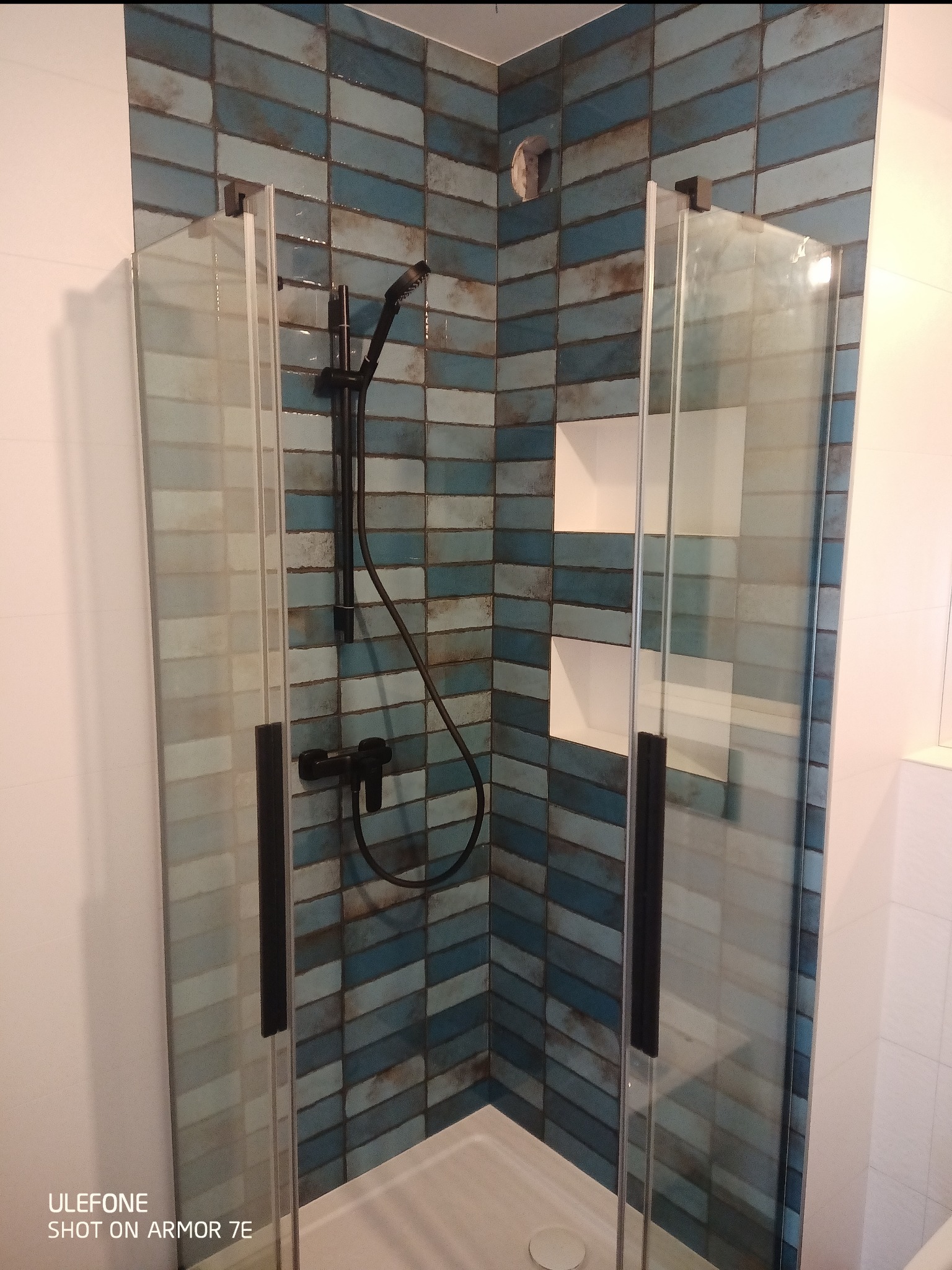 Kabina prysznicowa z niebiesko-szarą mozaiką ułożoną w jodełkę, czarna armatura i wnęki na kosmetyki.